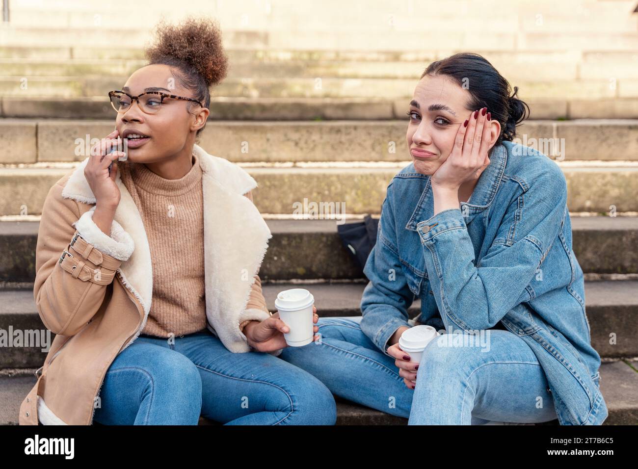 Zwei Studentinnen in einer Jeansjacke reden miteinander, am Telefon, lachen, trinken Kaffee auf der Treppe in der Stadt. Konzept der digitalen Teilung Stockfoto