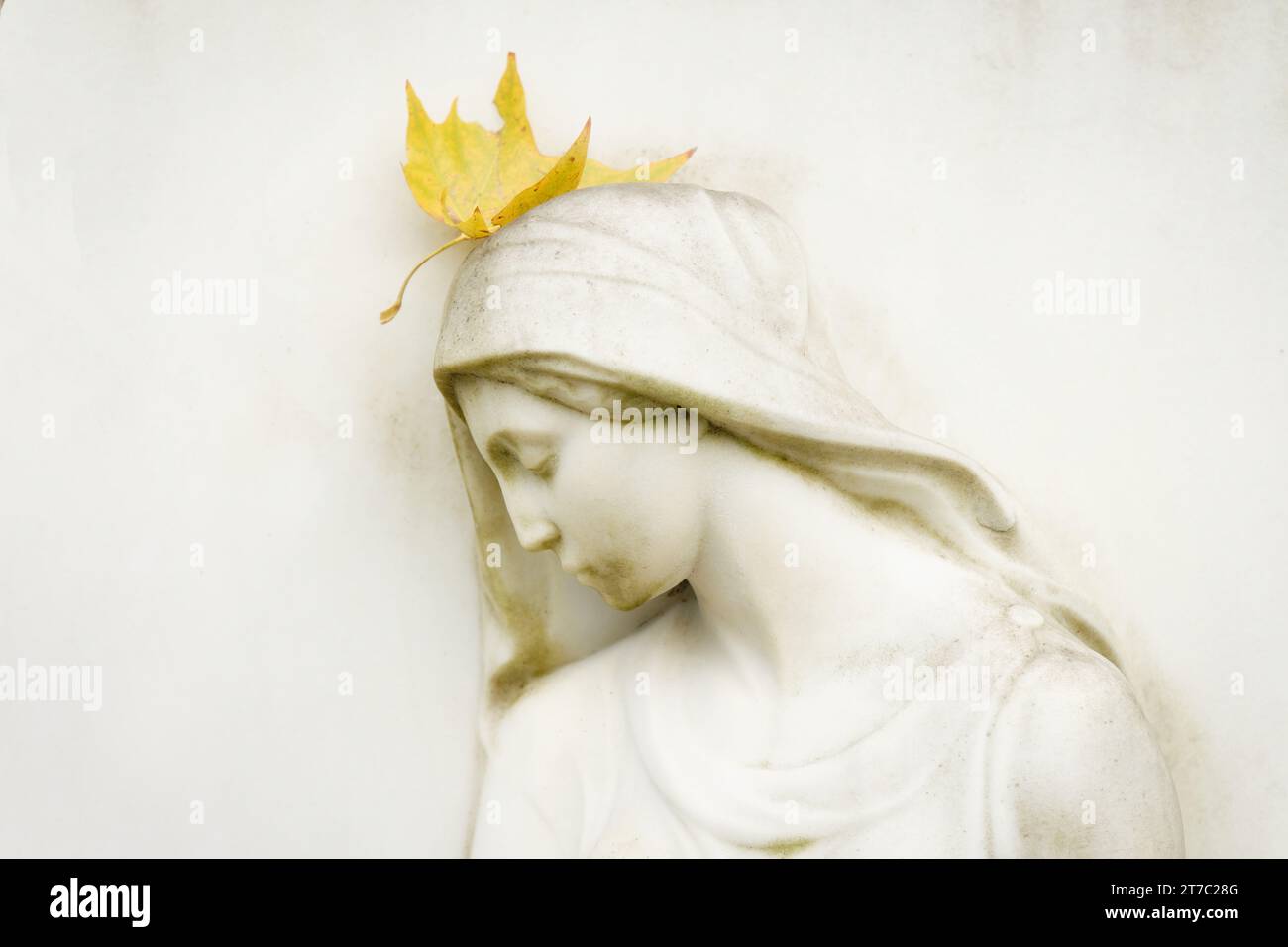 Die schöne weiße weibliche Figur auf einem Grabstein hat ein Herbstblatt auf dem Kopf wie eine Krone Stockfoto