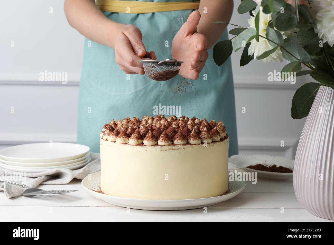 Frau, die leckeren Tiramisu-Kuchen mit Kakaopulver am weißen Holztisch bestäubt, Nahaufnahme Stockfoto