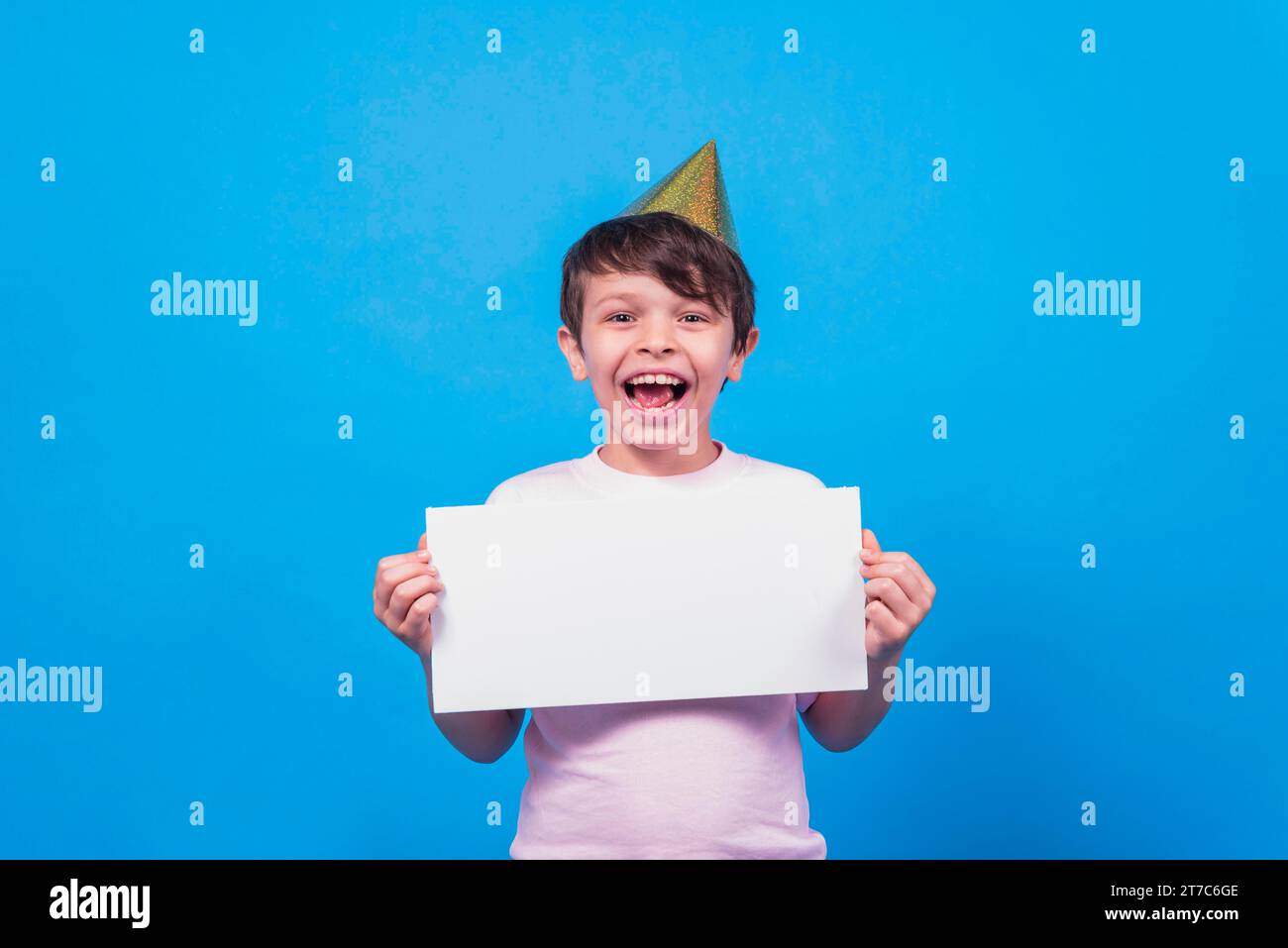 Aufgeregter kleiner Junge mit Partyhut, der leere Kartenhand mit blauer Oberfläche hält Stockfoto