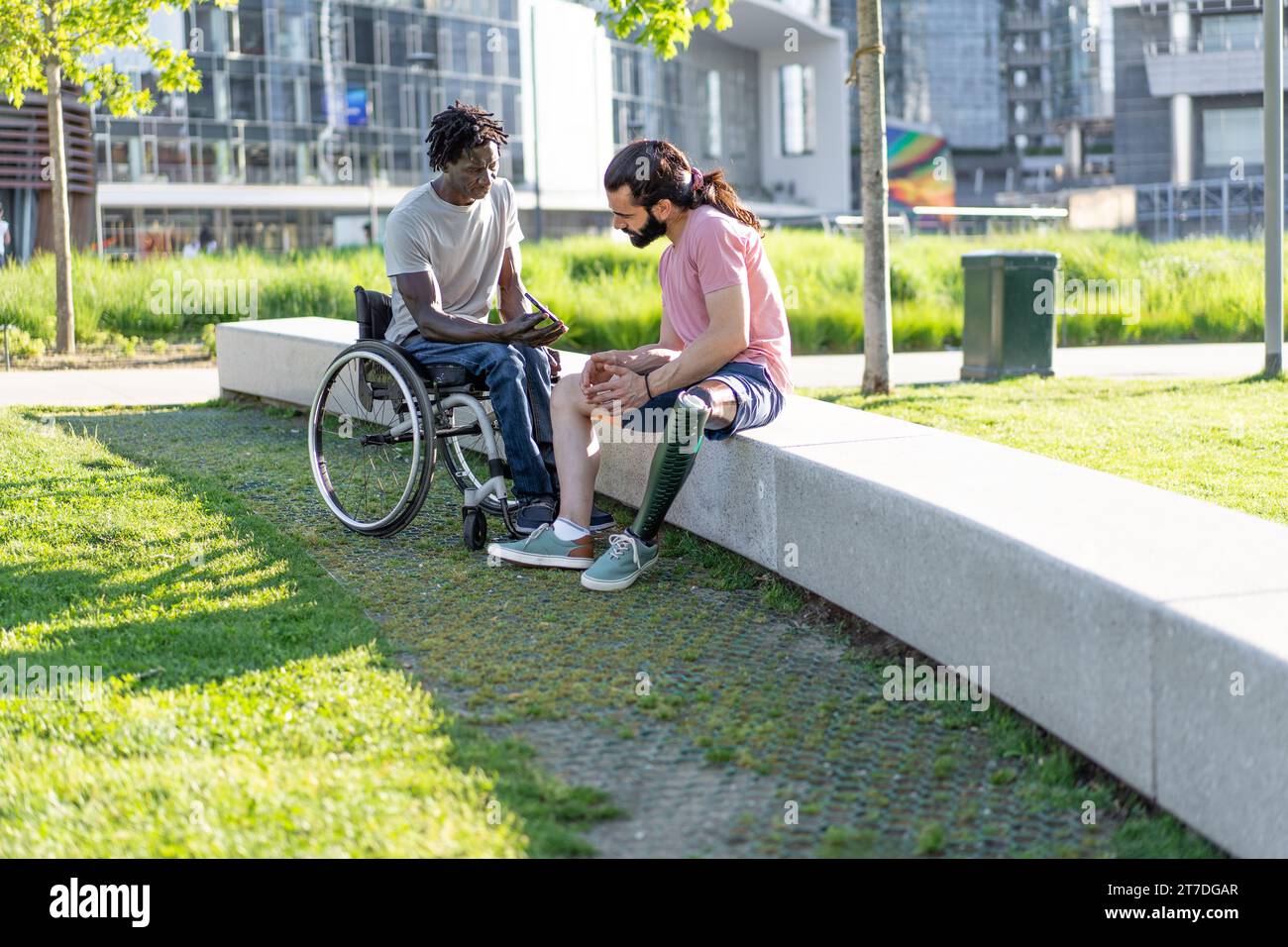Im Park unterhalten sich zwei Männer mit Inklusivität, einer auf einem Rollstuhl und der andere mit einer Beinprothese. Stockfoto
