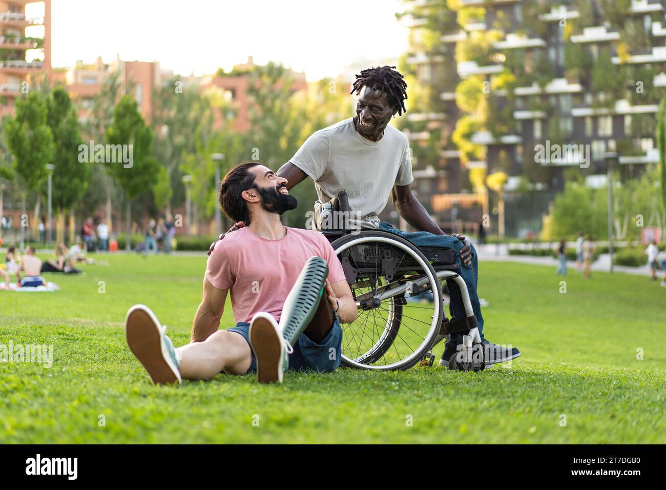 Zwei Männer, die im Park lachen, einer mit einer Beinprothese und der andere im Rollstuhl, genießen inklusiven Outdoor-Urlaub. Stockfoto
