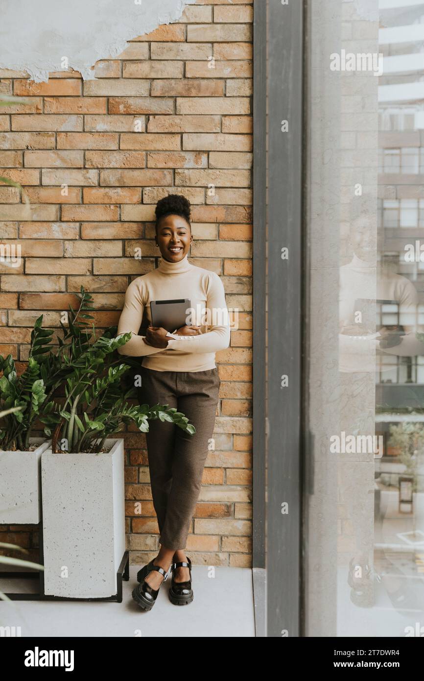 Eine hübsche junge afroamerikanische Geschäftsfrau mit digitalem Tablet, die an der Backsteinwand im Büro im Industriestil steht Stockfoto