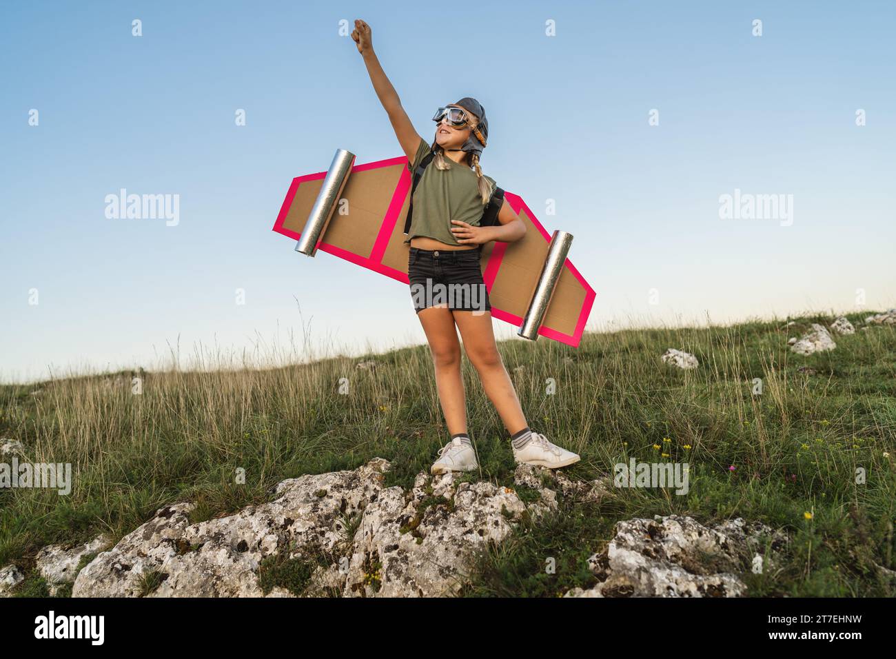 Überschwängliches Kind mit Pappflügeln und helmhebendem Arm im Triumph auf einem Felsen Stockfoto