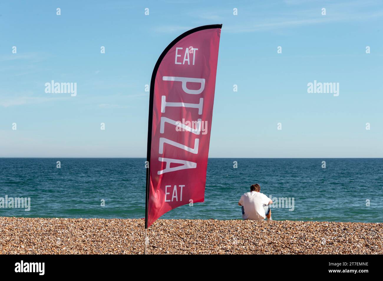 Essen Sie Pizza-Flagge am Strand mit der Rückansicht eines Mannes, der mit seiner Pizza zum Mitnehmen sitzt Stockfoto