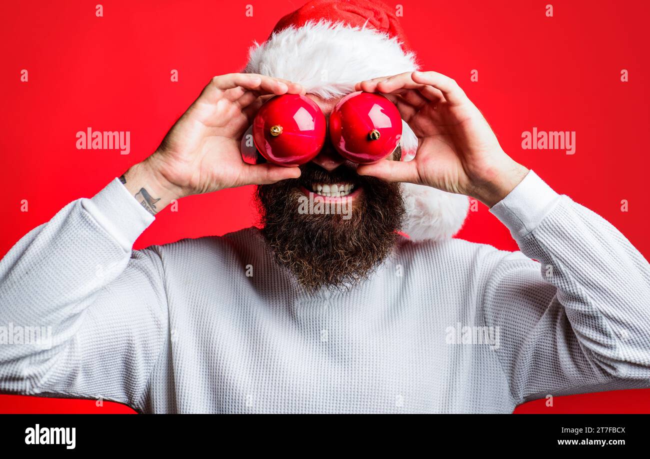 Bärtiger Mann mit Weihnachtsmann-Hut mit zwei Spielzeugen für Weihnachtsbaumaugen. Weihnachtsstimmung. Frohe Ferien. Weihnachtsmann mit dekorativen Weihnachtskugeln in der Nähe Stockfoto