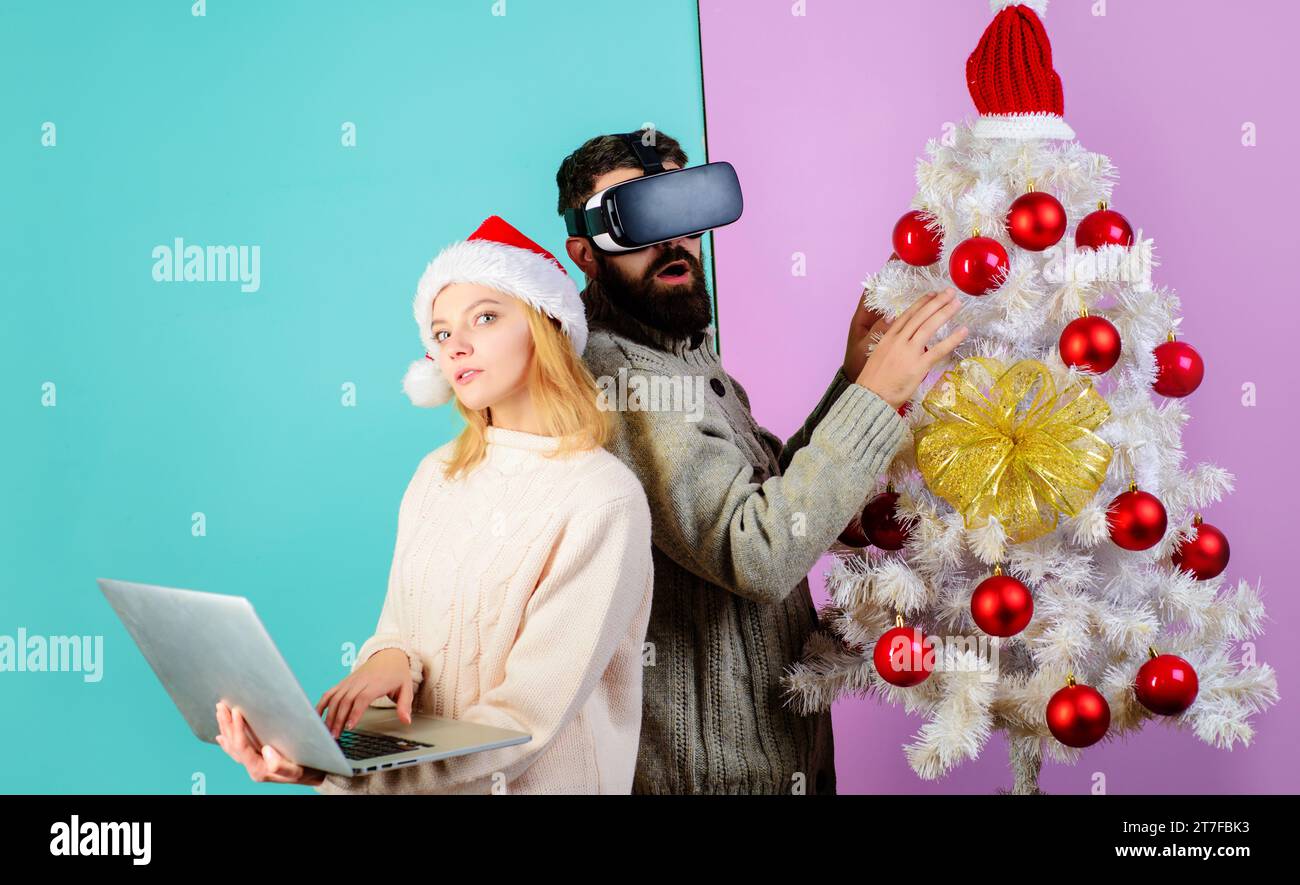 Weihnachtsfamilie mit VR-Brille und Laptop. Bärtiger Mann in Virtual-Reality-Brille-Headset. Frau mit Weihnachtsmann-Hut mit Notizbuch. Weihnachtspaar in der Nähe Stockfoto