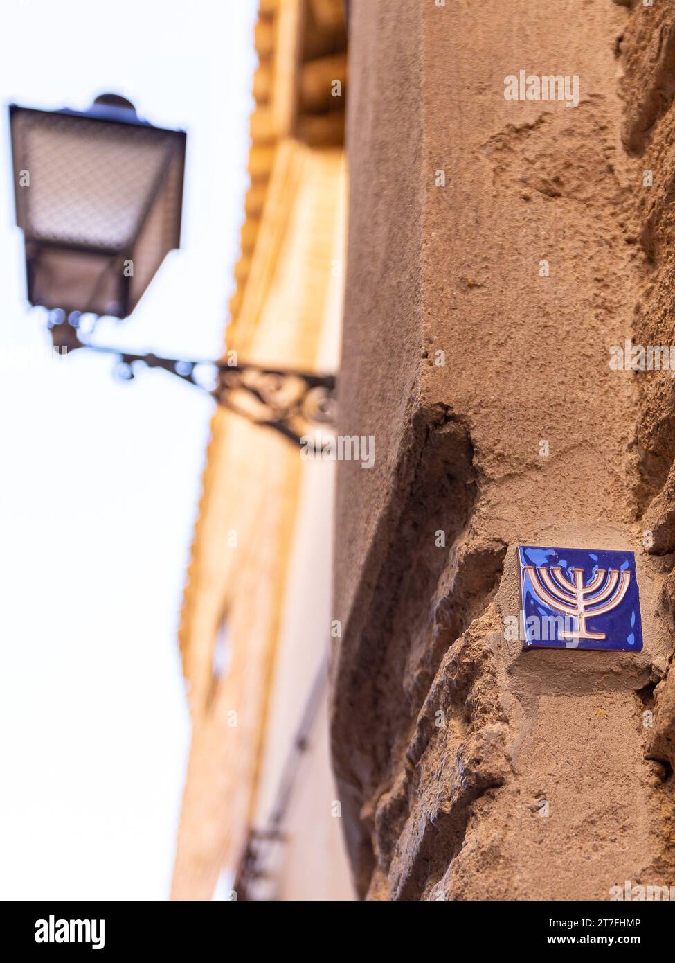 Kleine blaue Fliese mit Menora-Symbol, eingebettet in die Steinfassade im ehemaligen jüdischen Viertel in Toledo, Spanien, Straßenlaterne im alten Stil. Stockfoto