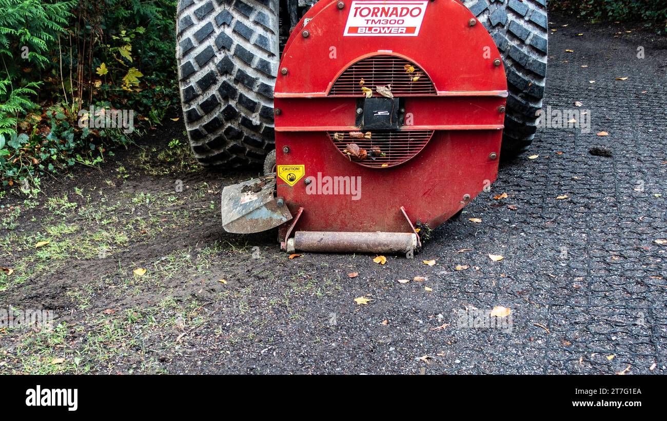 Nahaufnahme eines roten Tornado TM300 Laubbläsers, der an einem großen Fahrzeug befestigt ist und Herbstlaub von einem Parkweg entfernt, mit sichtbarem Warnschild. Stockfoto