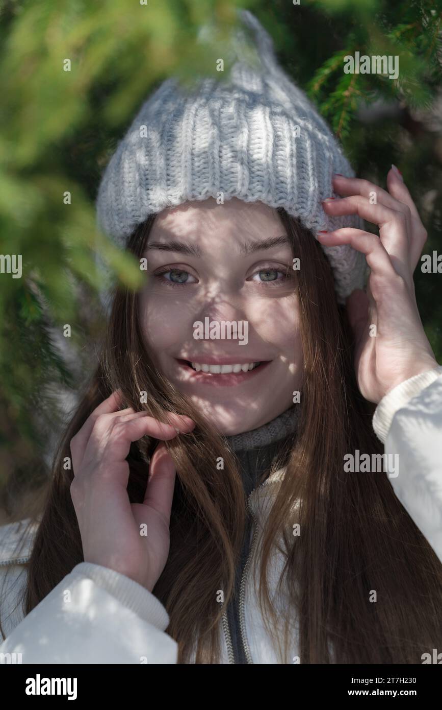 Porträt eines glücklichen Teenagers, der in die Kamera blickt, im Winterwald die Hände zu Gesicht erhob. Lächelnder Teenager mit langen braunen Haaren und ohne Make-up Stockfoto