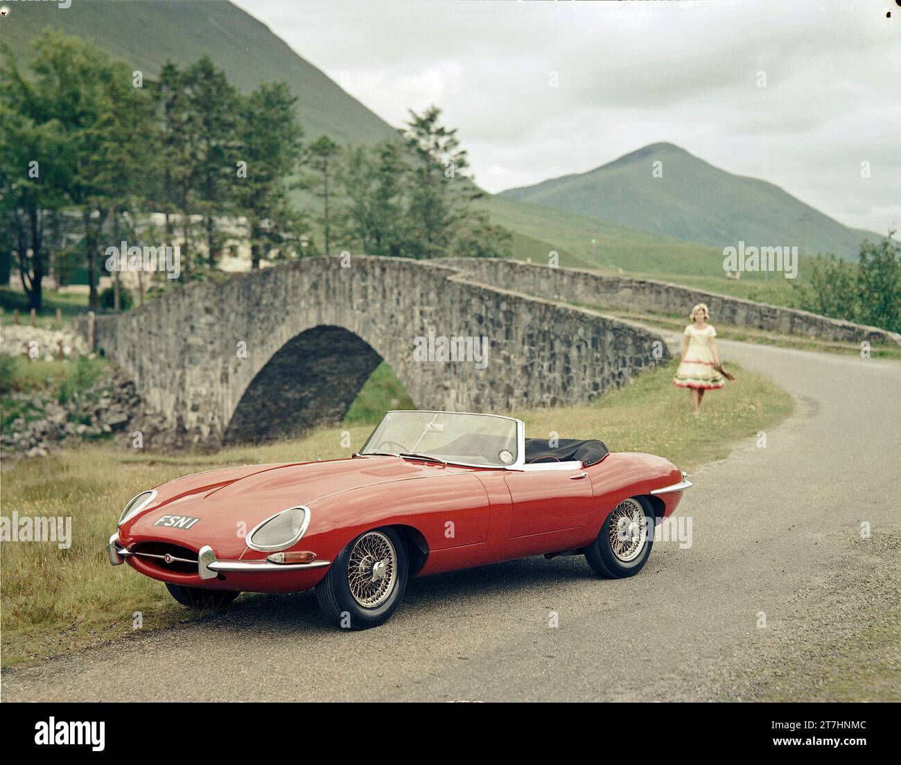 1961 Jaguar E-TYPE Roadster der Serie 1 neben einer Brücke in den schottischen Highlands mit weiblichem Modell in farbenfroher Kleidung Stockfoto