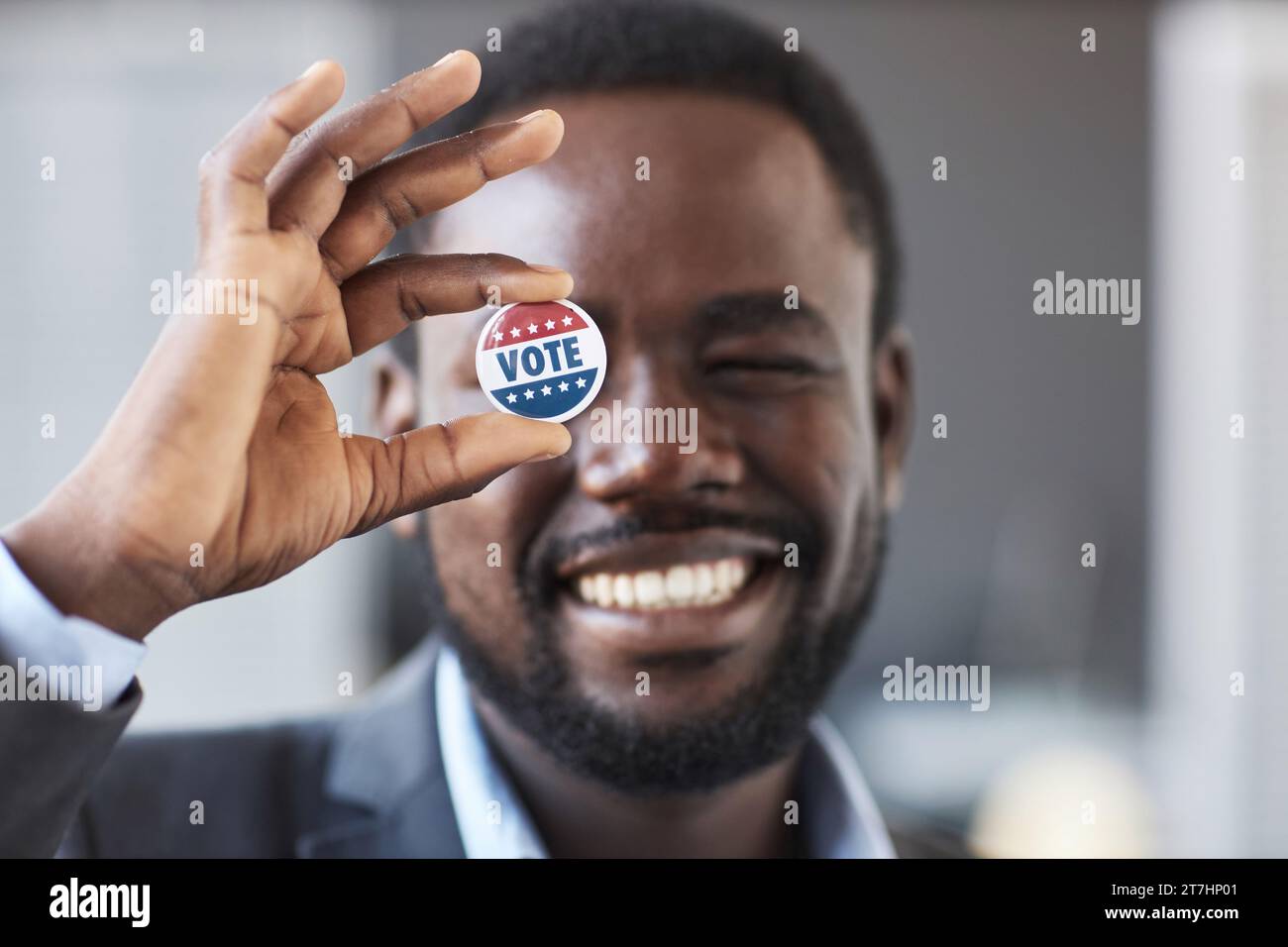 Konzentrieren Sie sich auf die Hand eines jungen, lächelnden afroamerikanischen Mannes, der das Wahlabzeichen vor seinem rechten Auge hält, während er vor der Kamera steht Stockfoto