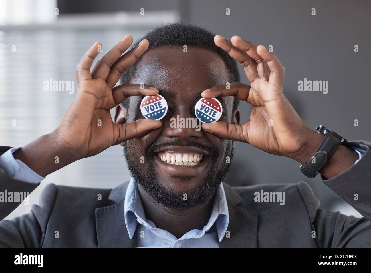 Glücklicher junger afroamerikanischer Wähler mit einem breiten, zahnlosen Lächeln, der zwei kleine runde Stimmabzeichen an den Augen hält, während er vor der Kamera steht Stockfoto