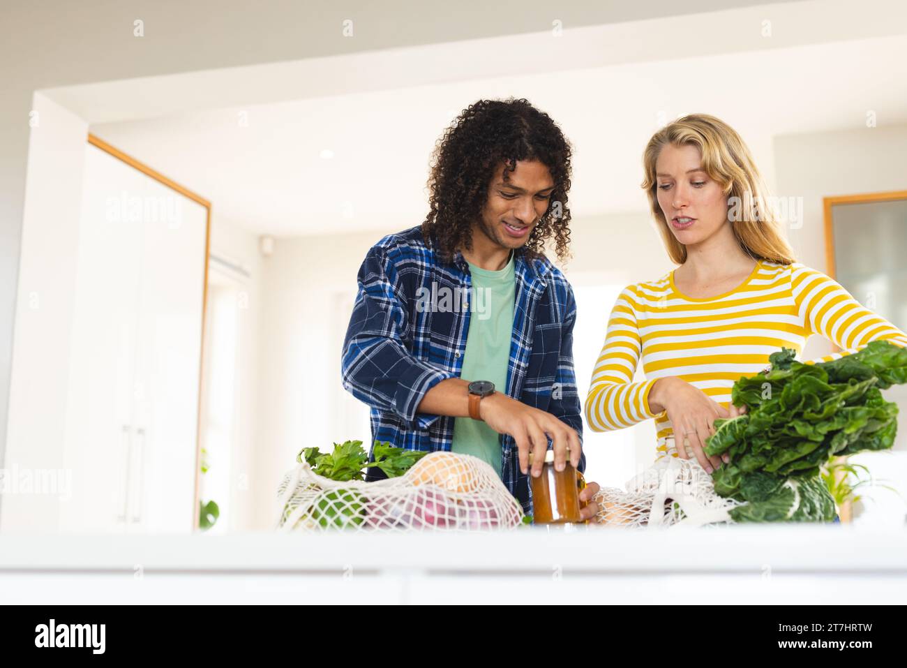 Glückliches, vielfältiges Paar, das Lebensmittel auspackt und in der sonnigen Küche diskutiert Stockfoto