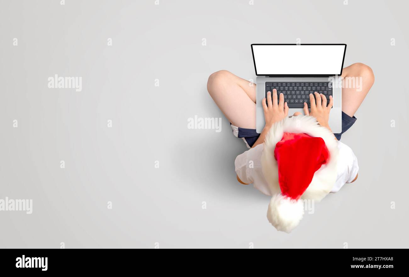 Junge in rotem Weihnachtsmütze arbeitet auf einem Laptop mit isoliertem Bildschirm für Mockup. Festliche Produktivität auf dem Boden. Kopierbereich Stockfoto