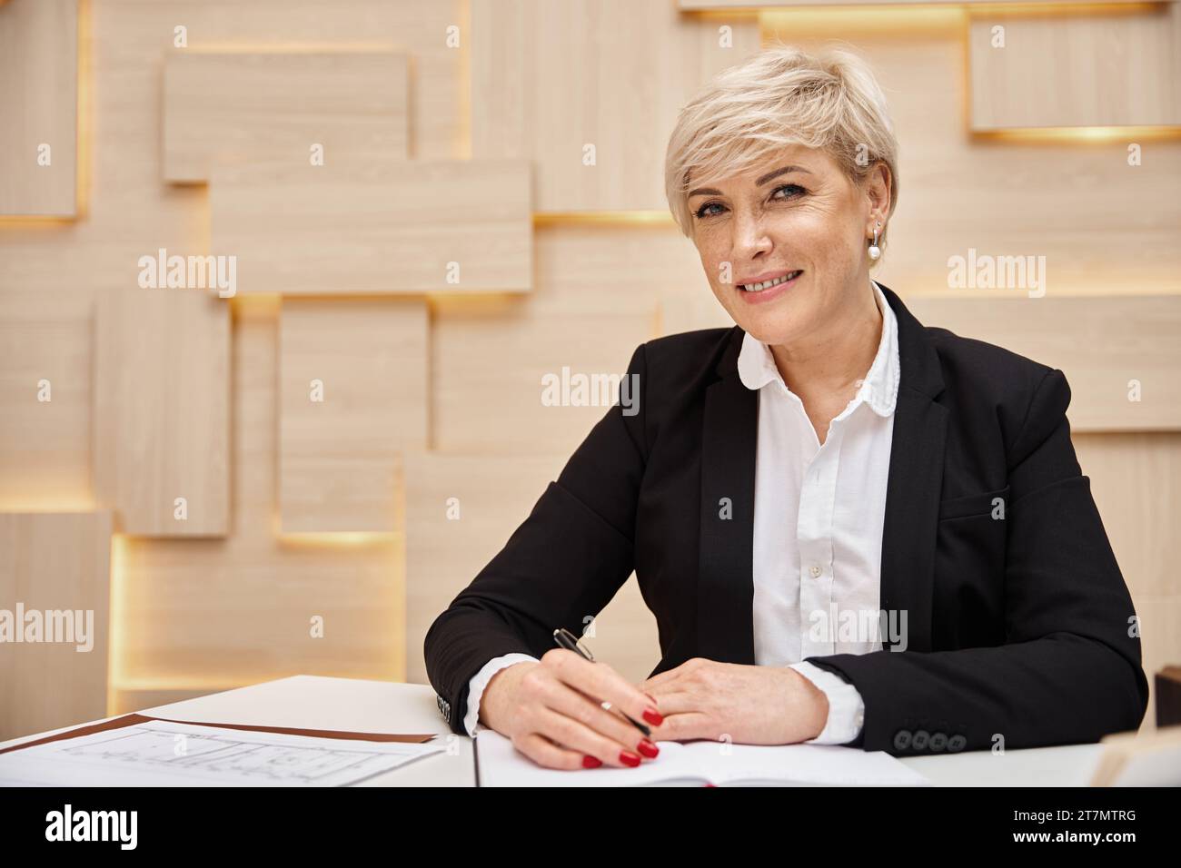 Glücklicher blonder Immobilienmakler mittleren Alters mit kurzen Haaren, der im Immobilienbüro arbeitet und in die Kamera schaut Stockfoto