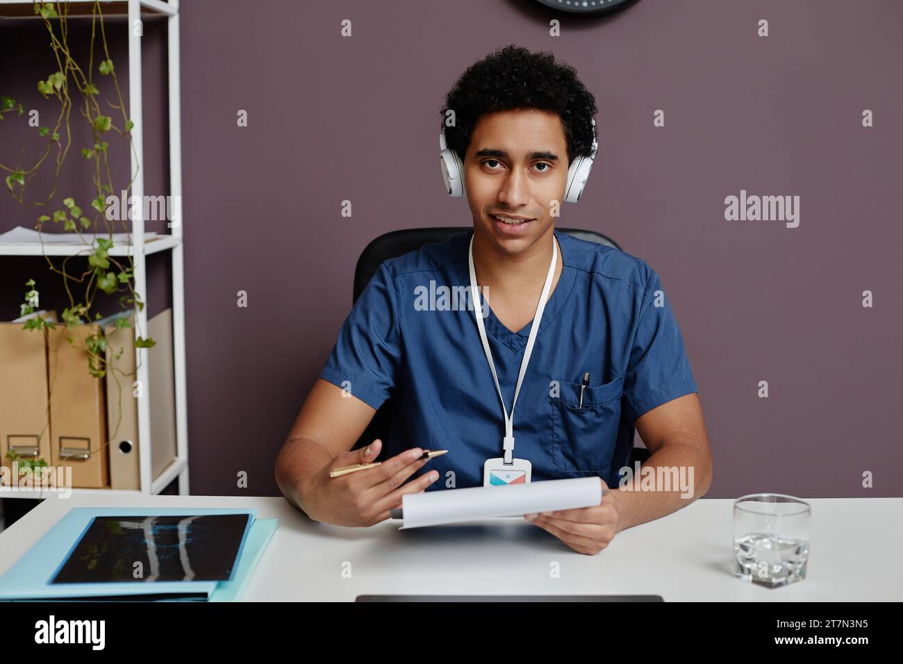 Vorderansicht des jungen arabischen Mannes als Arzt, der Kopfhörer mit Geräuschminimierung am Arbeitsplatz trägt und mit dem Patienten spricht, Kopierraum Stockfoto