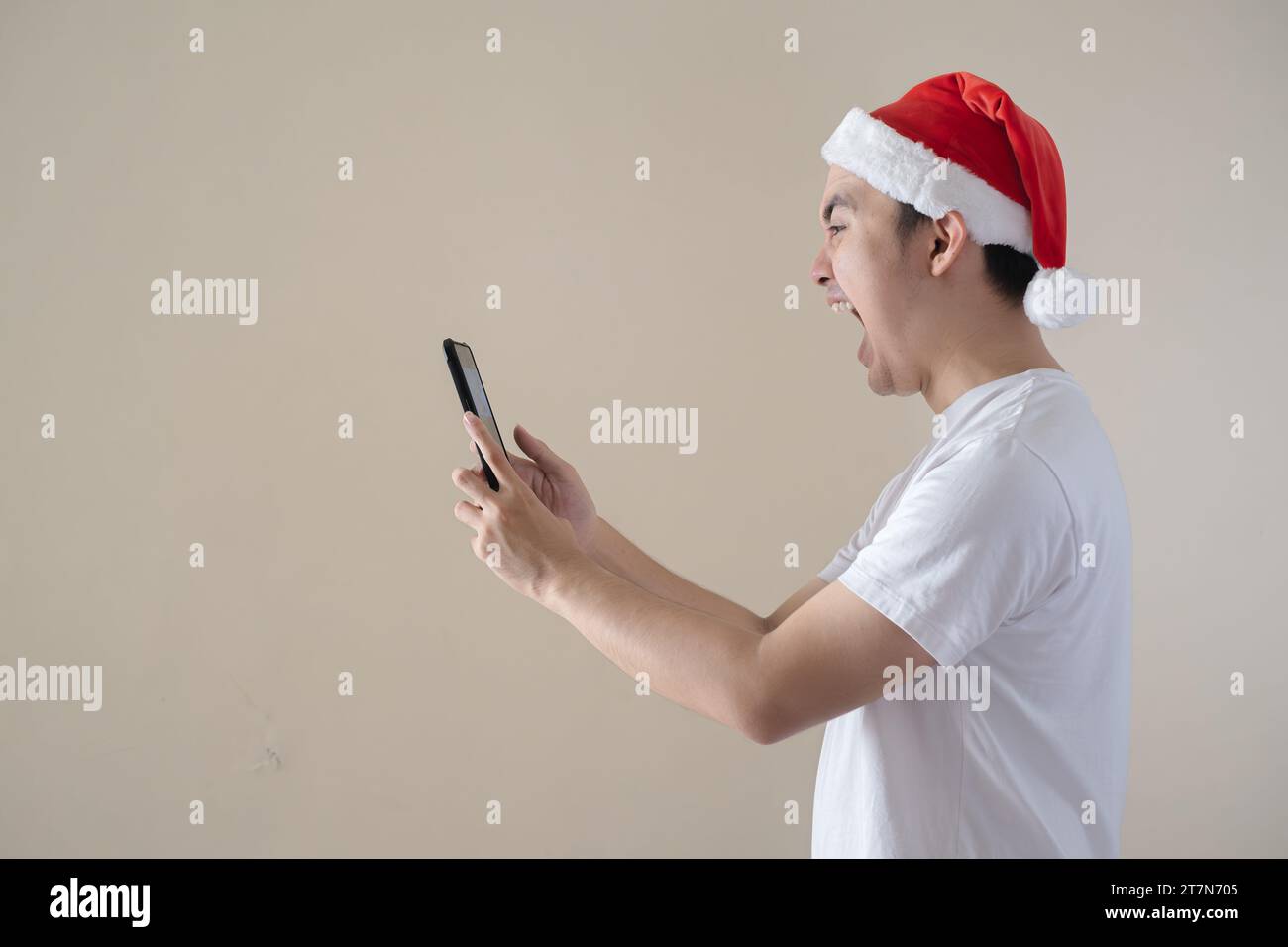 Der junge Weihnachtsmann ist wütend, während er am Telefon schaut. Isolierter beigefarbener Hintergrund. Weihnachtskonzept. Stockfoto