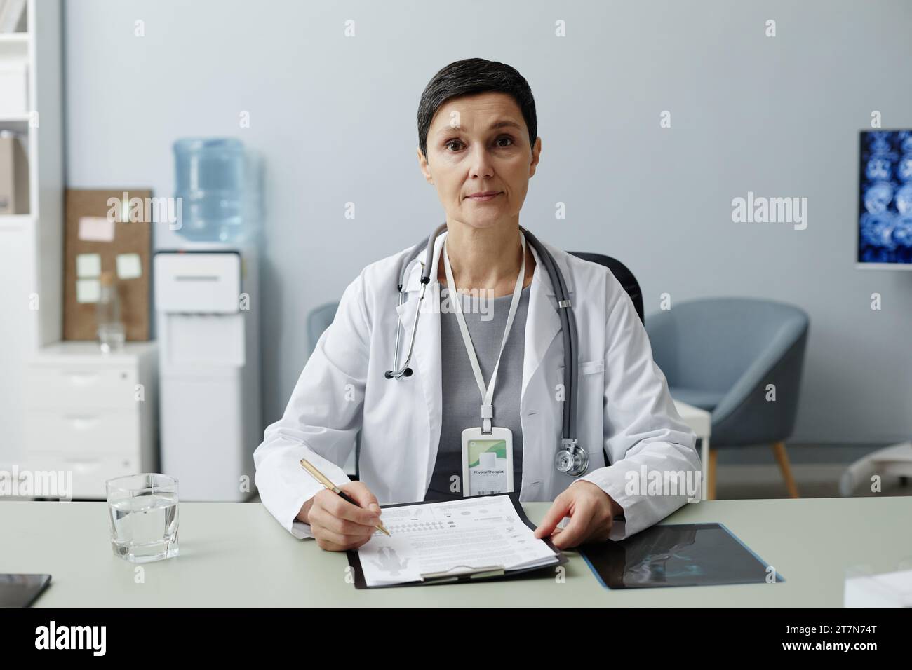 Frontporträt der Reifen Frau als Ärztin, die am Arbeitsplatz in der Klinik sitzt und in die Kamera blickt Stockfoto