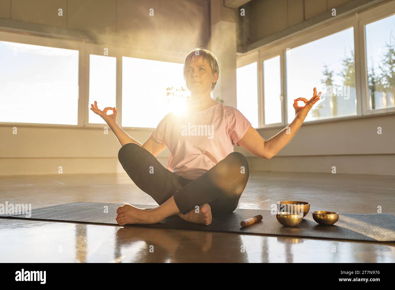 Frau in Meditationspose mit Klangschalen, ruhiger Fitnessraum-Atmosphäre, Sonnenschein Stockfoto