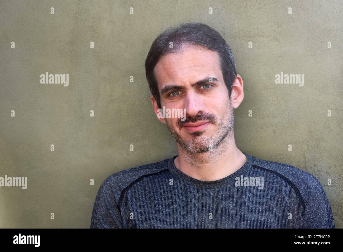 Außenporträt eines jungen kaukasischen 30-jährigen Mannes mit kurzem Bart, schwarzem Haar und blauen Augen, der vor einer flachen grünen Wand in die Kamera lächelt. Spa kopieren Stockfoto