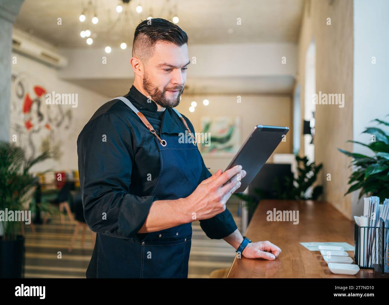 Lächelnder Inhaber eines kleinen Unternehmens in schwarzer Chefuniform mit Schürze und digitalem Tablet in seinem gemütlichen Restaurantsaal. Erfolgreiche Menschen, hart W Stockfoto