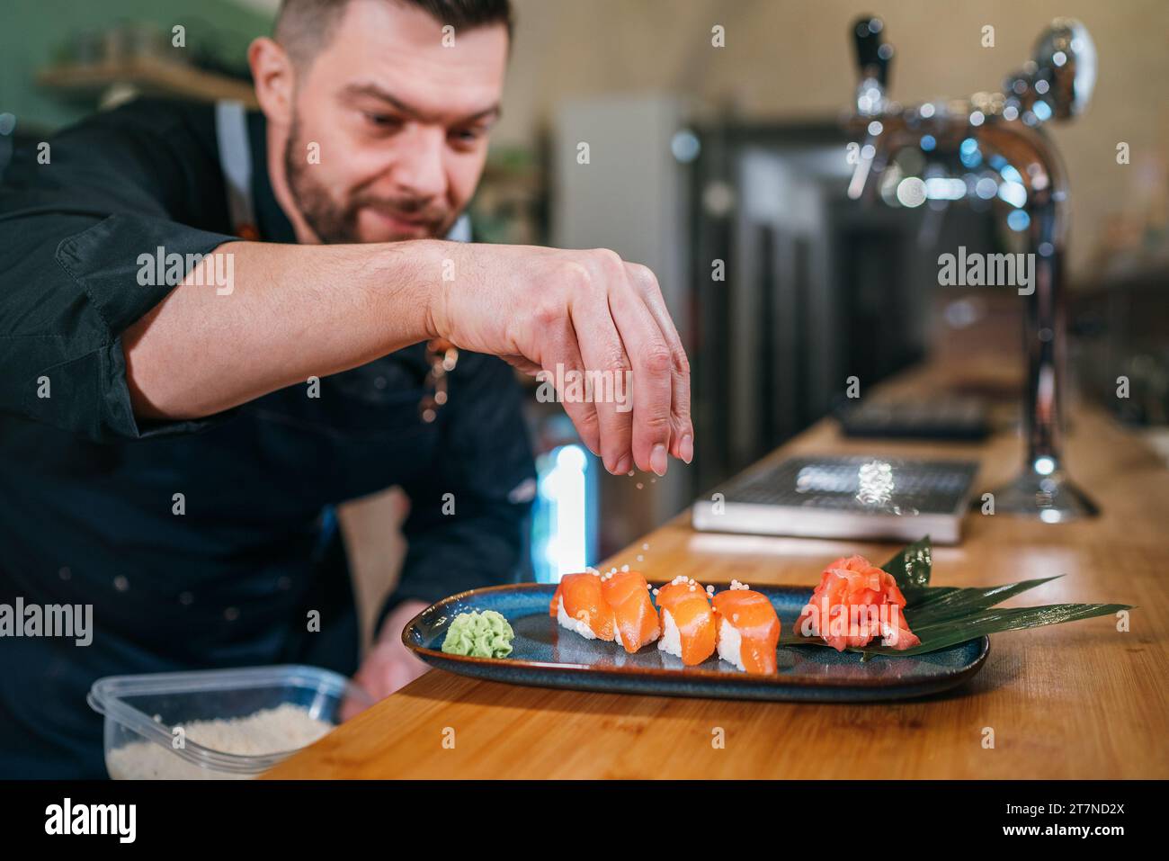 Der bärtige Koch kocht in schwarzer Uniform und bestreut das Sushi Nigirizushi Gericht in der Küche des Restaurants im Keller. Berufsberuf, Lebensmittelvorbereitung Stockfoto