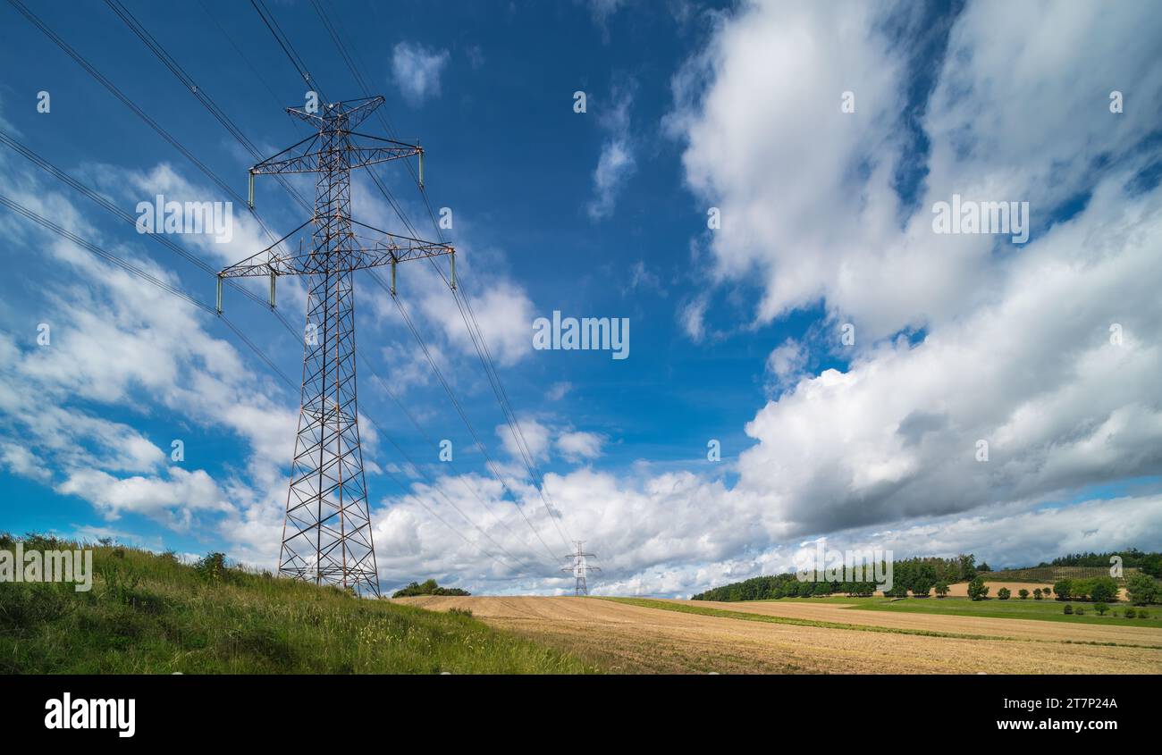 Oberleitungsmast in der Sommerlandschaft. Elektrischer Traversen-Übertragungsturm mit Hochspannungsdrähten, grünem Gras, Stoppeln, Feld oder Bäumen am Berg. Stockfoto