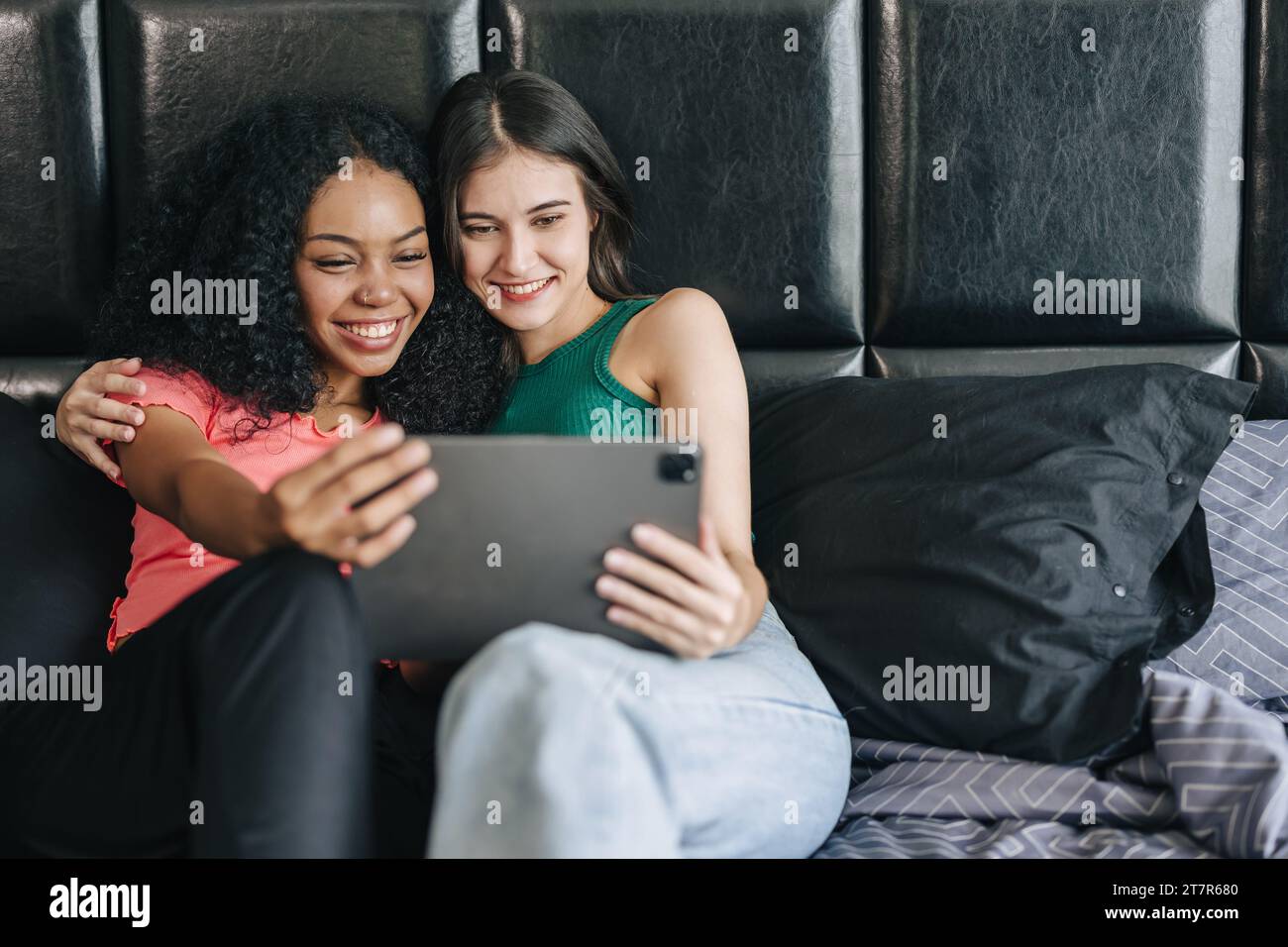 LGBT-Frauen, die auf dem Bett sitzen und einen Tablet-Bildschirm sehen, glücklich lächelnd, zusammen zu Hause, Urlaub Aktivität Stockfoto