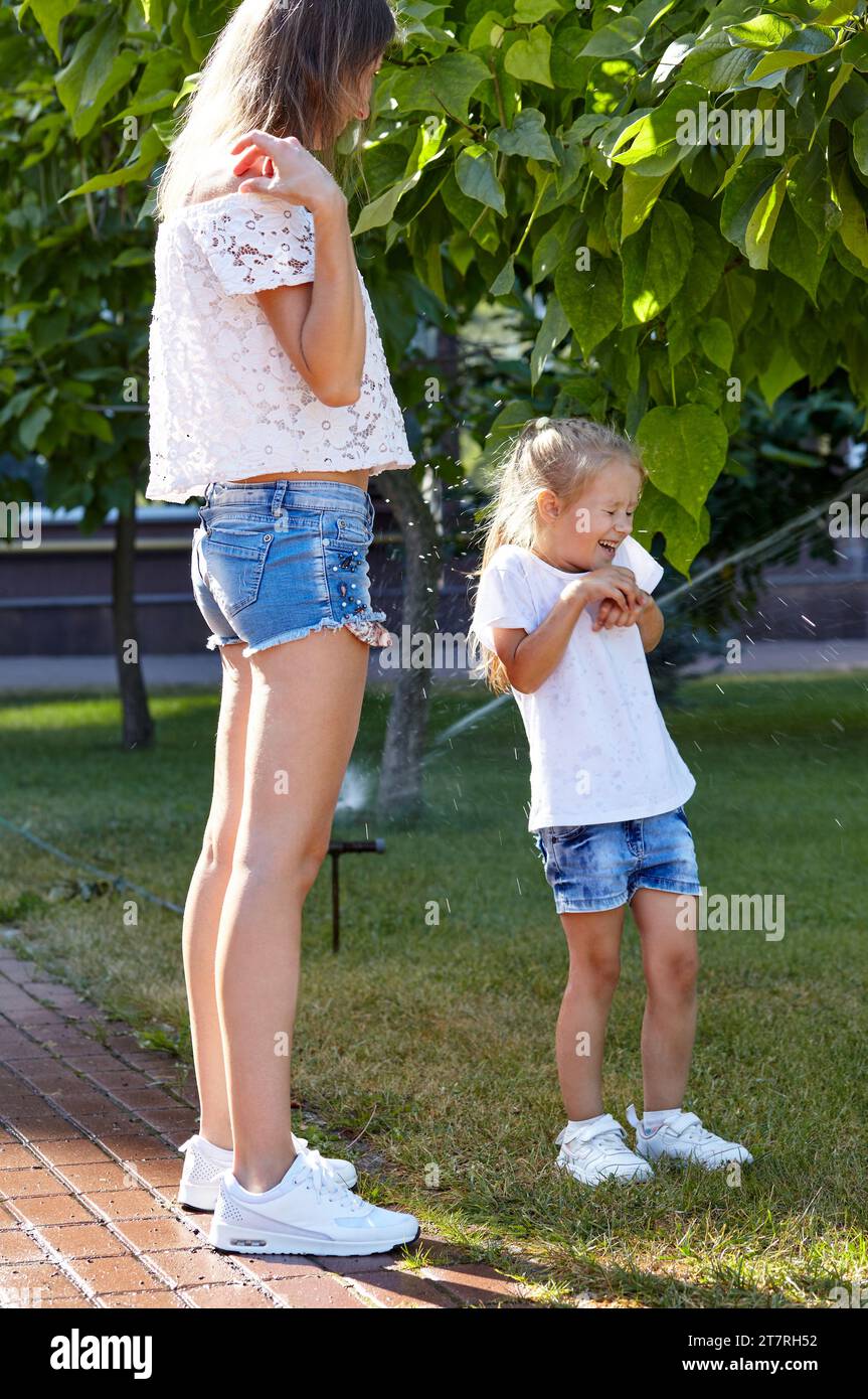 Mutter mit Tochter, die unter dem Wasser von der Sprinklerbewässerung im Sommer Stadtpark läuft. Kindheit, Freizeit und Menschenkonzept - Happy fami Stockfoto