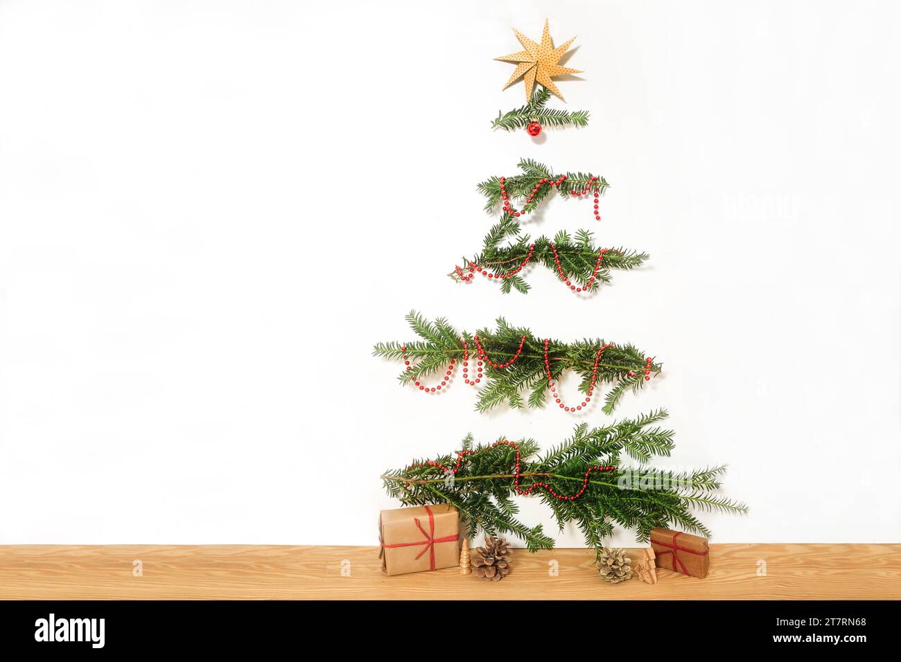 DIY-Weihnachtsbaum aus natürlichen Zweigen, die an eine weiße Wand genagelt sind, rote Dekorationskette und ein Papierstern auf der Oberseite, Geschenke auf dem Holzboden, einfach Stockfoto