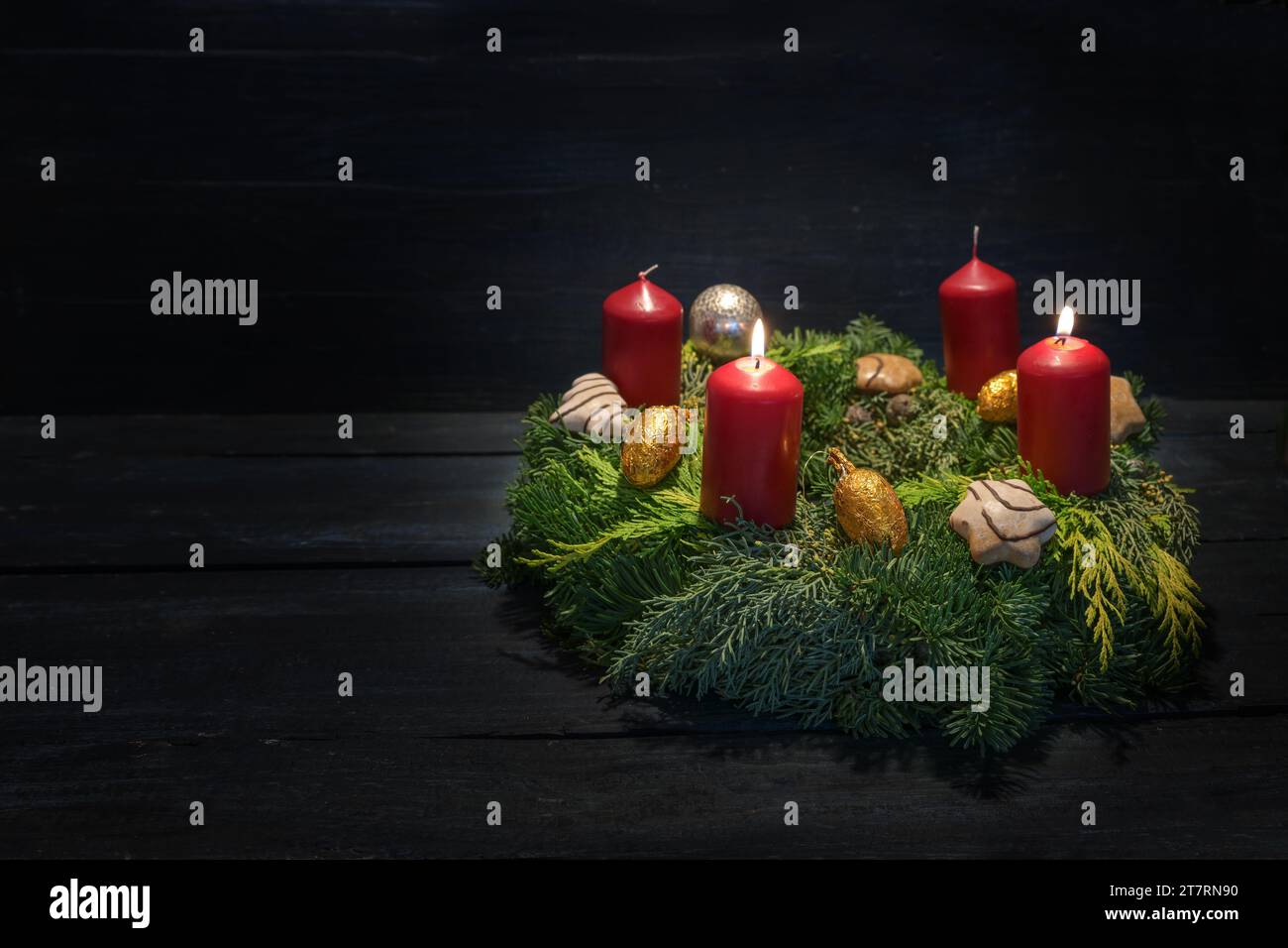 Licht im Dunkeln am zweiten Advent, naturgrüner Kranz mit roten Kerzen, zwei brennen, Weihnachtsdekoration und Kekse, dunkler hölzerner Hintergrund, Stockfoto