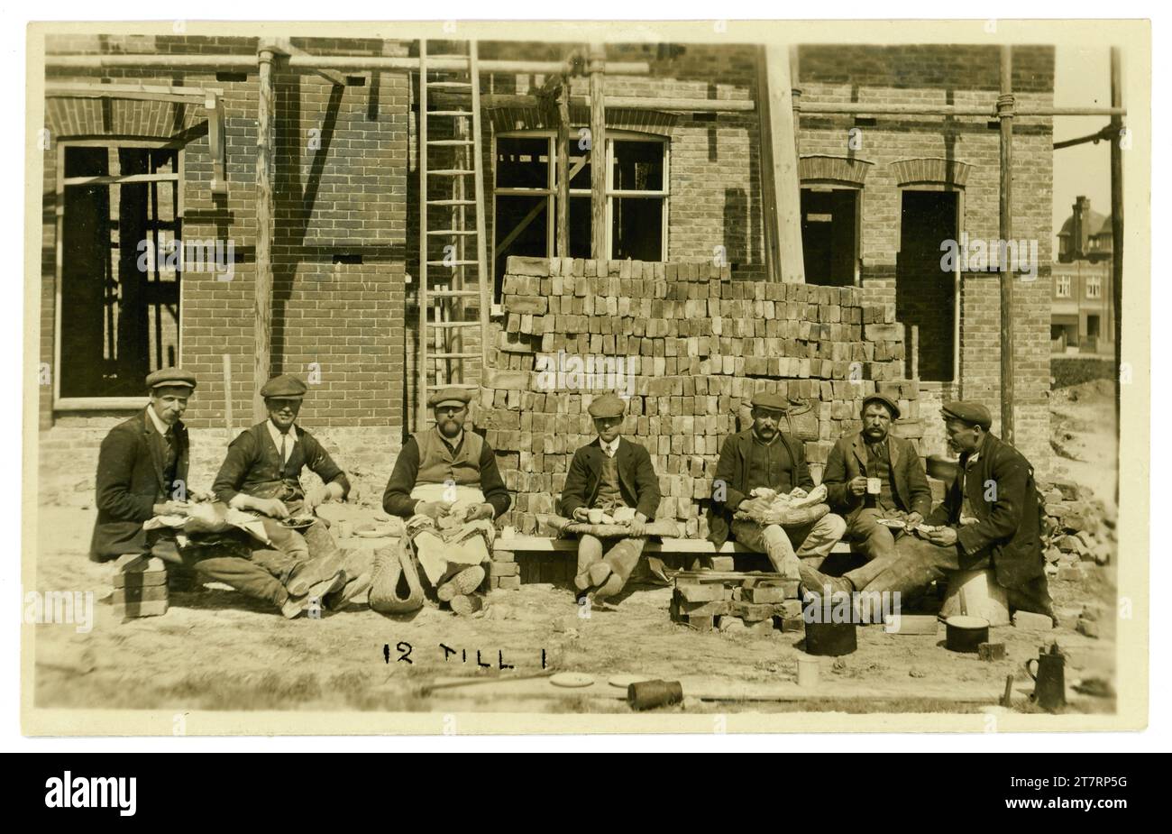 Originalpostkarte einer entspannten Gruppe von Maurern, die während ihrer Mittagspause vor einem Haus, das noch im Bau ist, Mittagessen essen, Anfang der 1920er Jahre von Kragen datiert, auf der Vorderseite steht eine Notiz mit der Aufschrift 12 bis 1. Einige gute Charaktere. GROSSBRITANNIEN Stockfoto