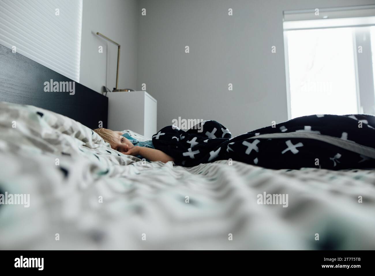 Weite Seitenansicht eines kleinen Jungen, der in einem großen Bett liegt und seinen Daumen lutscht Stockfoto