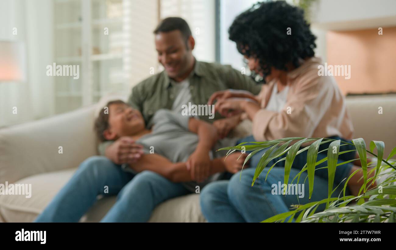 Familie afroamerikanische Eltern mit einem kleinen adoptierten Sohn verbinden, der auf dem Sofa kitzelt und zusammen aufrichtig lacht und Spaß hat Stockfoto