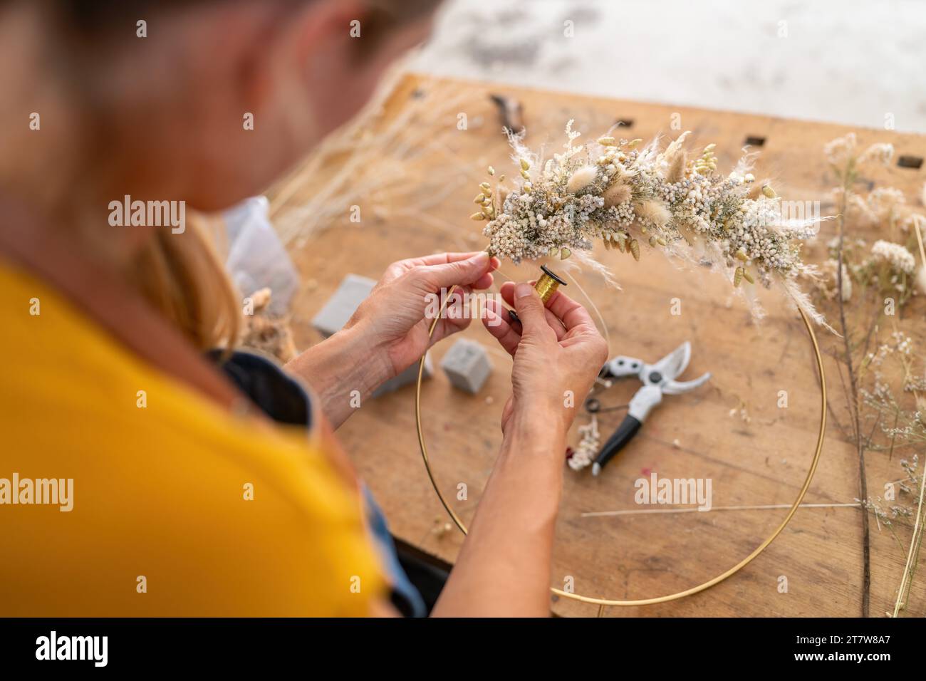 Nahaufnahme der Hände eines Floristen, die getrocknete Blumen an einem Ring vor einem hölzernen Tischhintergrund befestigen Stockfoto
