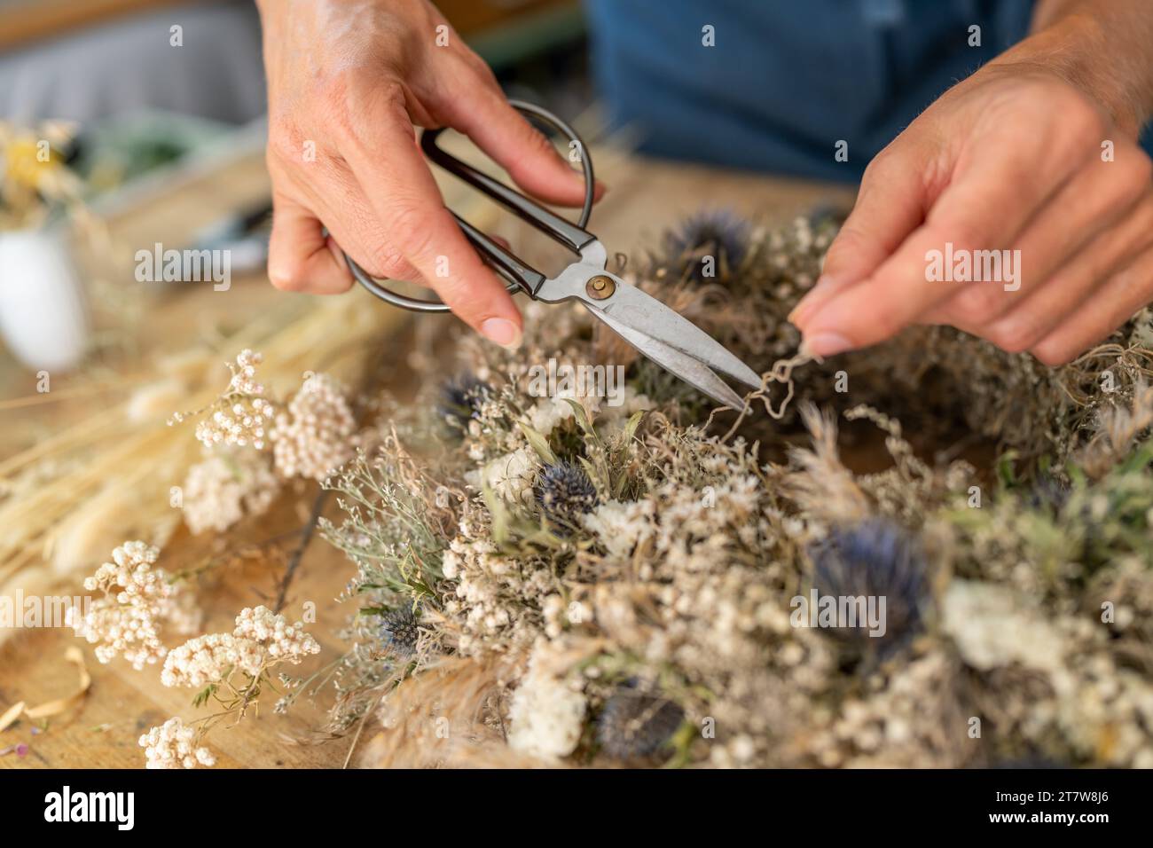 Hände mit einer Schere einen getrockneten Blumenkranz auf einem hölzernen Werkstatttisch zuschneiden Stockfoto