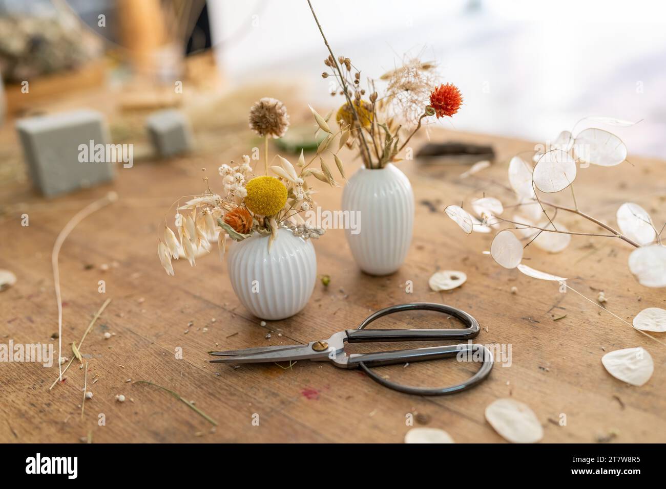 Zwei weiße gerippte Vasen mit getrockneten Blumen auf einem Holztisch mit verstreuten Blättern und Schere Stockfoto