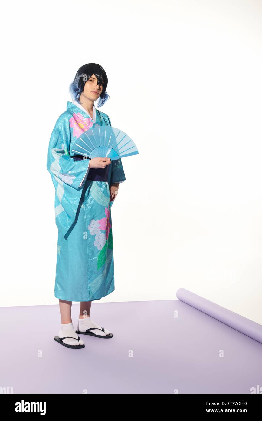 Junger Mann in blauem Kimono und Perücke posiert mit Handfächer auf lila Teppich und weißem Hintergrund, Cosplay Stockfoto