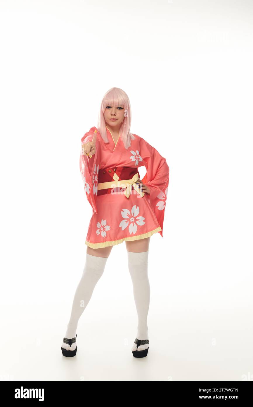 Die ganze Länge einer Frau in blonder Perücke und rosa Kimono, die auf die Kamera zeigt, auf weiße Cosplay-Kultur Stockfoto