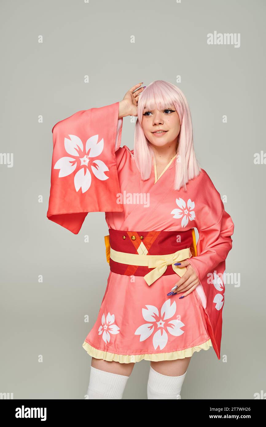 Junge lächelnde Frau in blonder Perücke und rosa Kimono mit Blumenmuster, die auf grauem Anime-Stil schweben Stockfoto