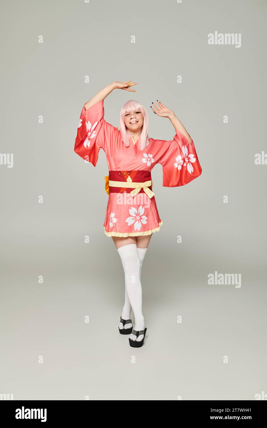 Cosplay blonde Frau in farbenfrohem Kimono und weißen Kniestrümpfen posiert mit erhobenen Händen auf Grau Stockfoto