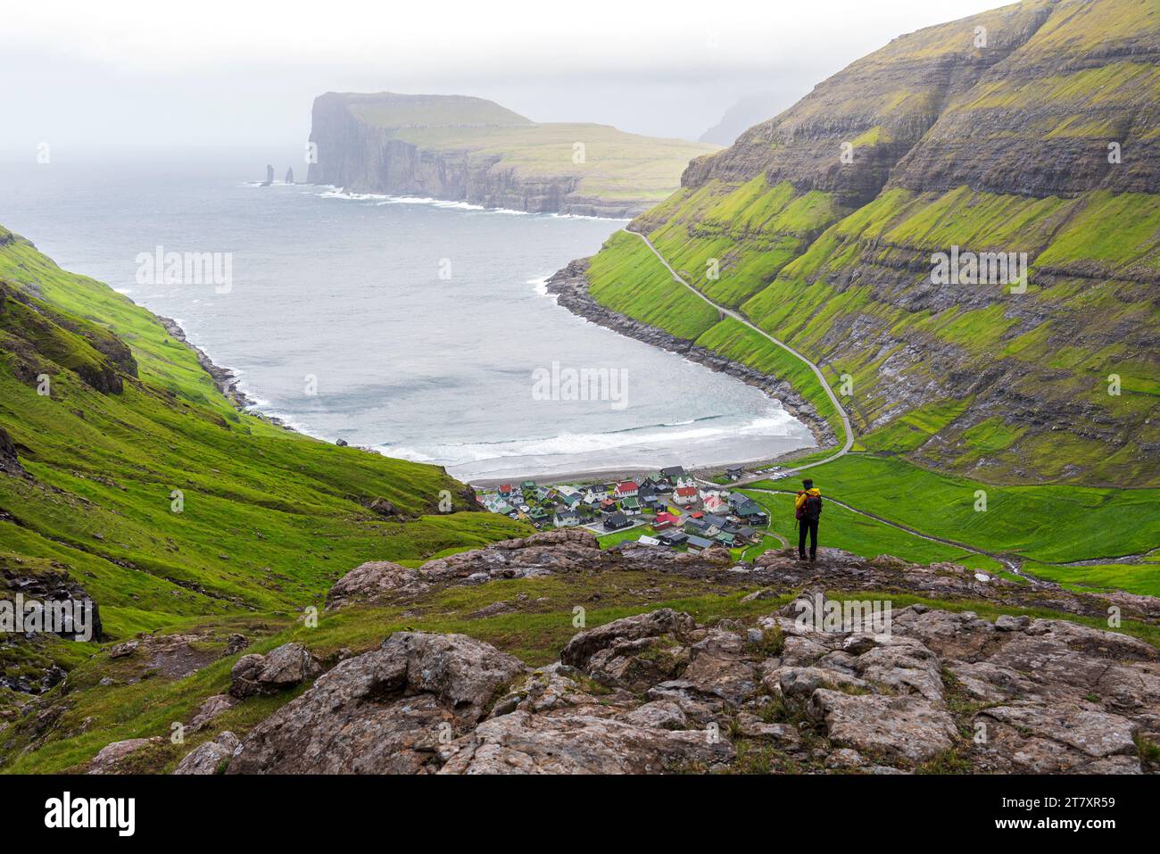 Ein Mann auf einem Felsen, der das Dorf Tjornuvik bei Nebel bewundert, Gemeinde Sunda, Insel Streymoy, Färöer, Dänemark, Europa Stockfoto