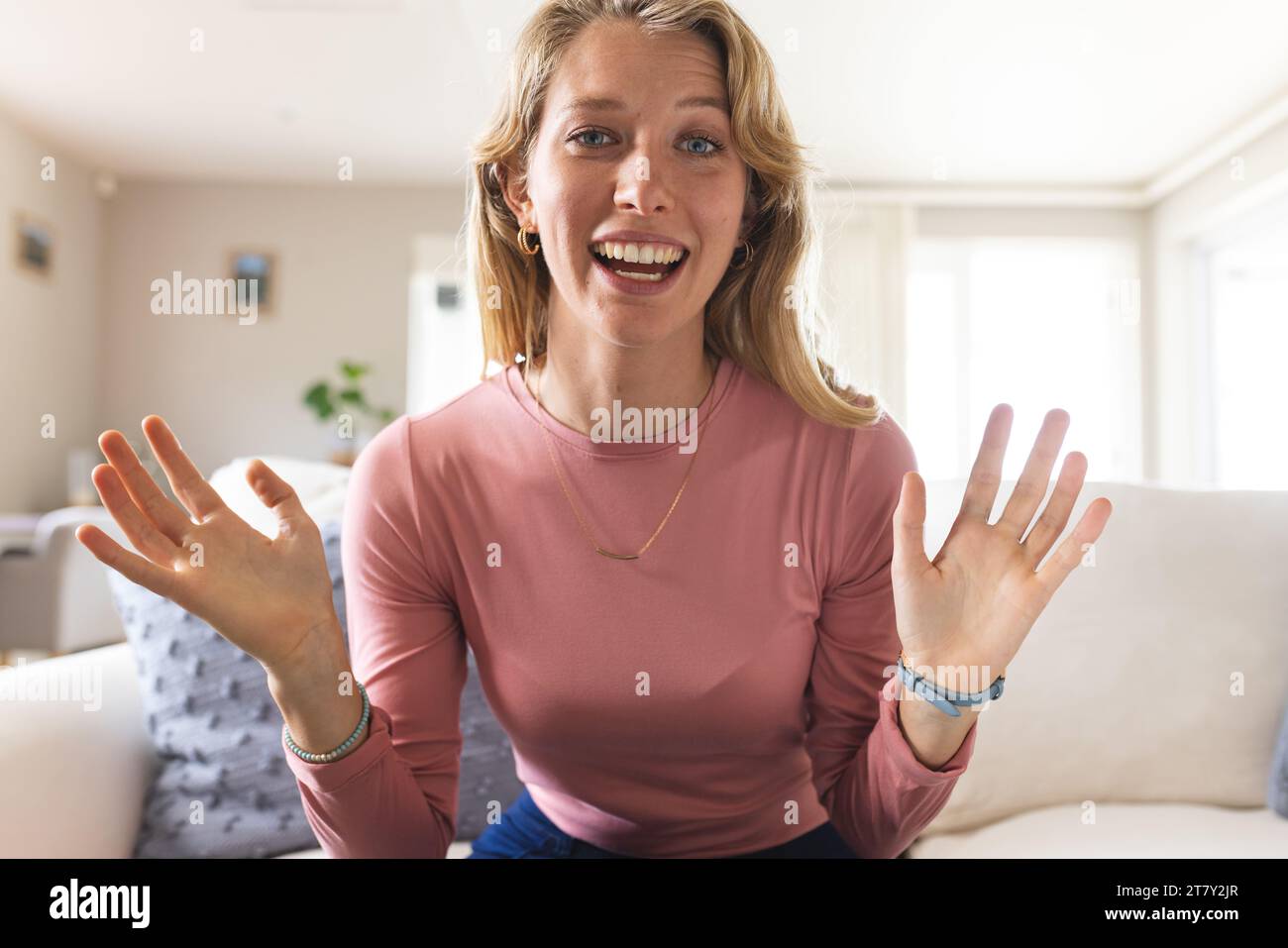 Glückliche blonde weiße Frau mit Videoanruf und Gestikulierung im sonnigen Wohnzimmer Stockfoto