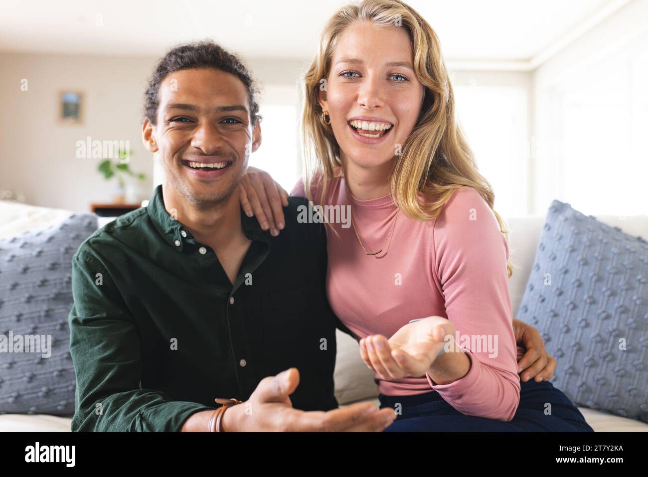 Glückliches, vielseitiges Paar mit Videoanruf und gestikuliertem Gesicht im sonnigen Wohnzimmer Stockfoto