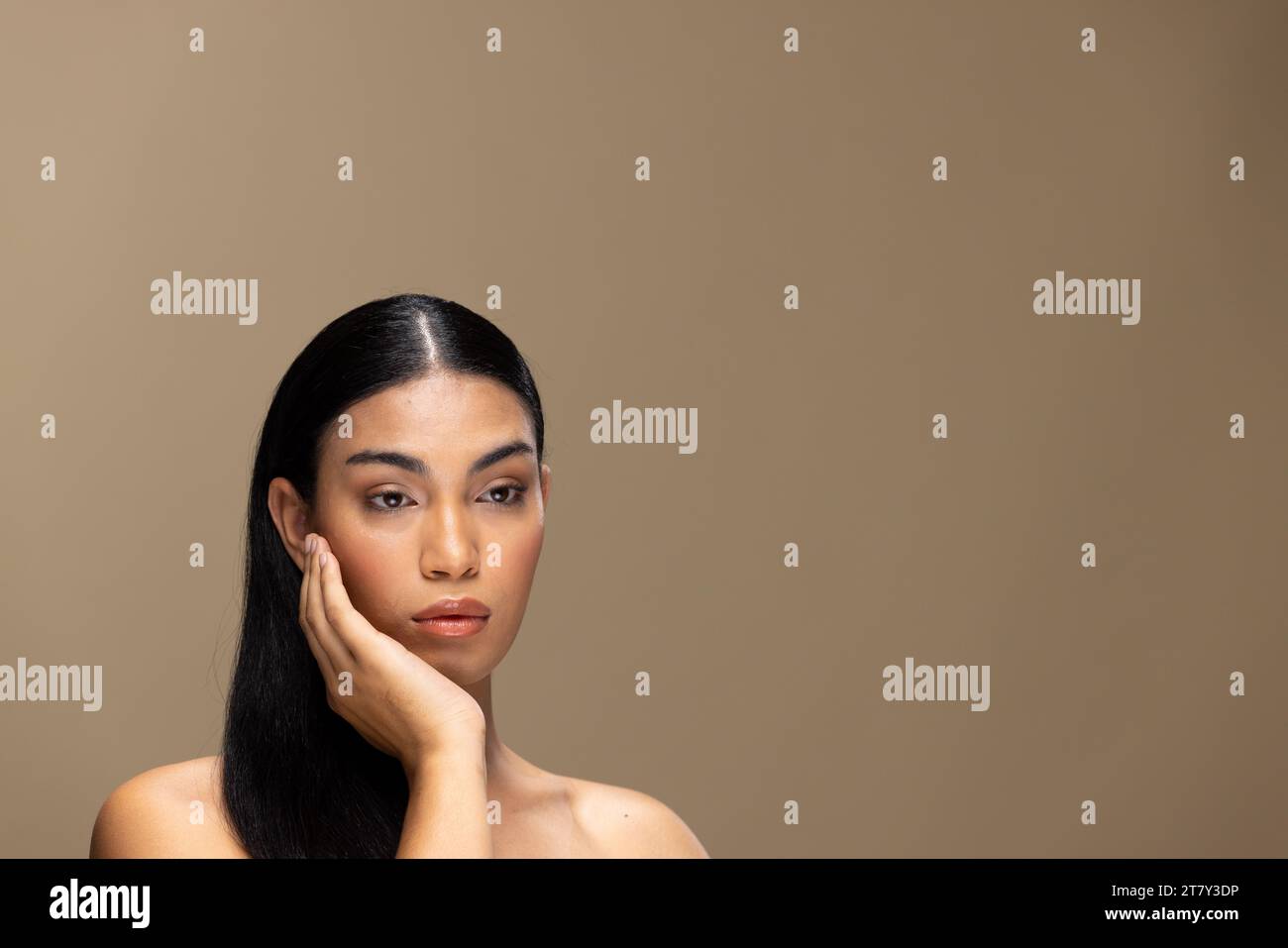 Porträt einer zweirassigen Frau mit dunklem Haar, Hand-zu-Gesicht und natürlichem Make-up auf braunem Hintergrund Stockfoto