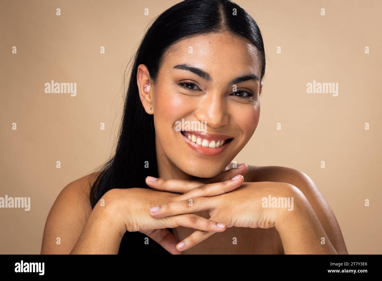 Lächelnde birassische Frau mit dunklen Haaren, Händen unter dem Kinn, natürliches Make-up auf braunem Hintergrund Stockfoto