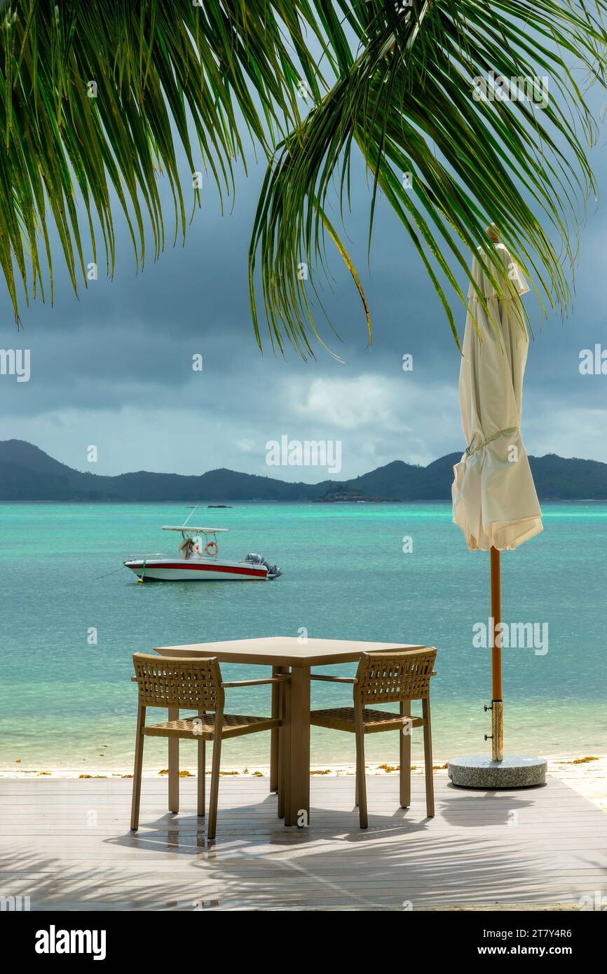 Leerer Tisch und Sonnenschirm unter einer Palme am Strand, Praslin Island, Frühstück oder Mittagessen in einem Strandrestaurant mit Aussicht, Seychellen Stockfoto