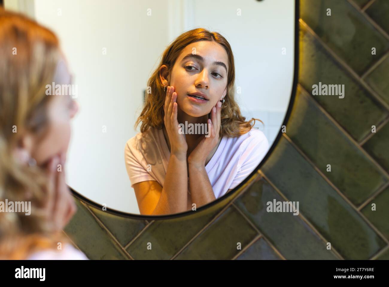 Glückliche birassische Frau, die das Gesicht berührt und zu Hause auf die Reflexion im Badezimmerspiegel blickt Stockfoto