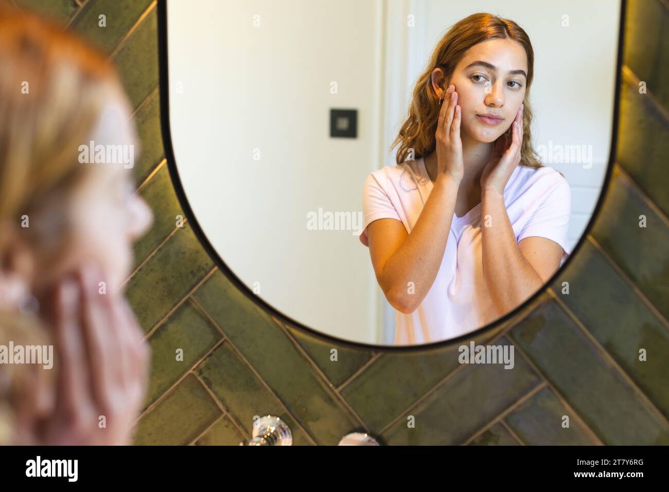 Glückliche birassische Frau, die das Gesicht berührt und zu Hause auf die Reflexion im Badezimmerspiegel blickt Stockfoto